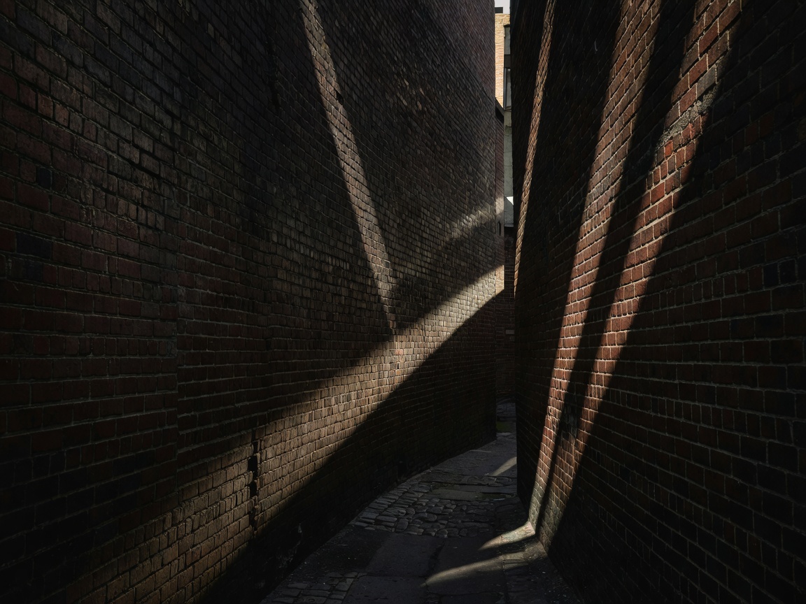 Enge Stadtgasse mit strukturierten Backsteinwänden, durchzogen von scharfen diagonalen Schatten